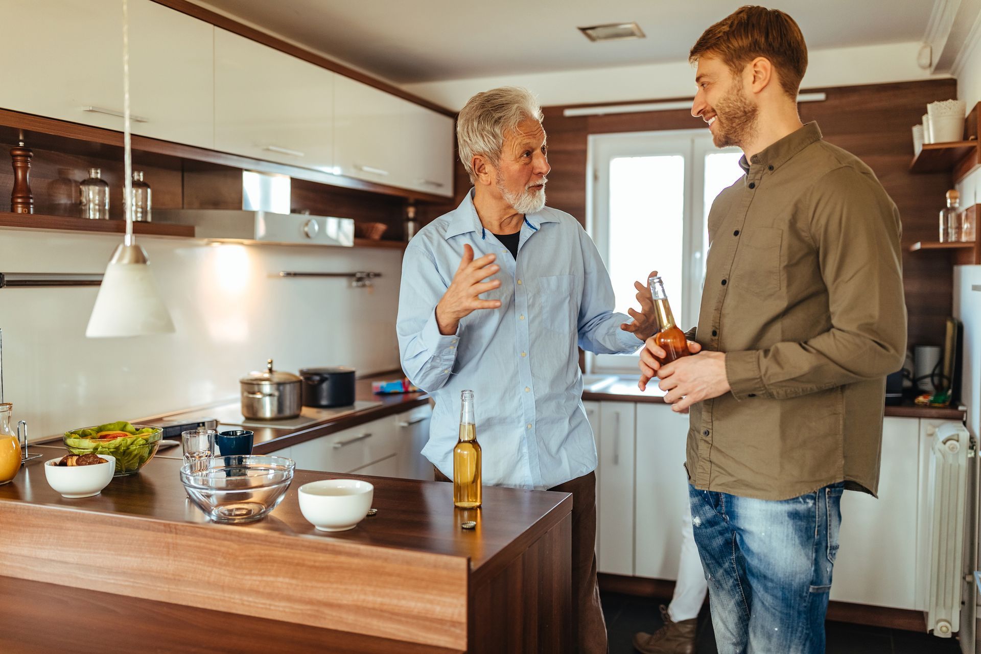 Two people stand in a modern kitchen, holding glass bottles and talking, with an island and cookware in the background.