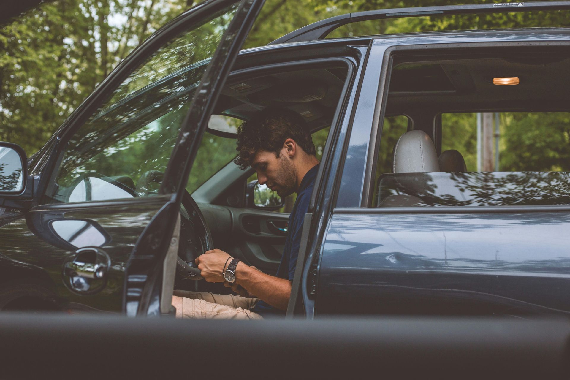 A person sits in the driver’s seat of a dark SUV with the door open, looking down at their hands while wearing a watch.