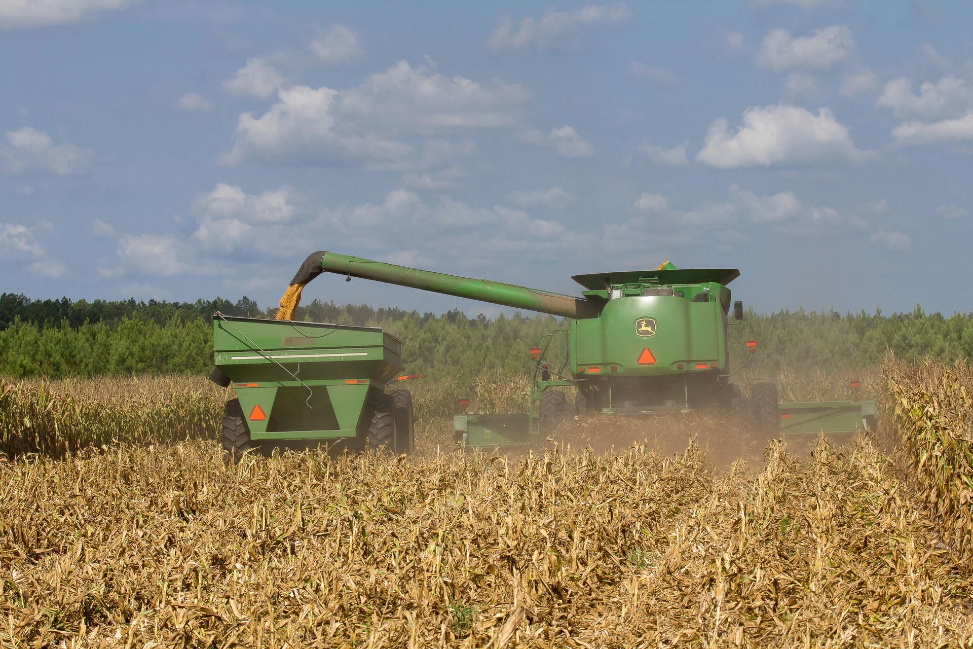 A green combine harvester offloads harvested corn into a grain cart while moving through a field under a cloudy sky.