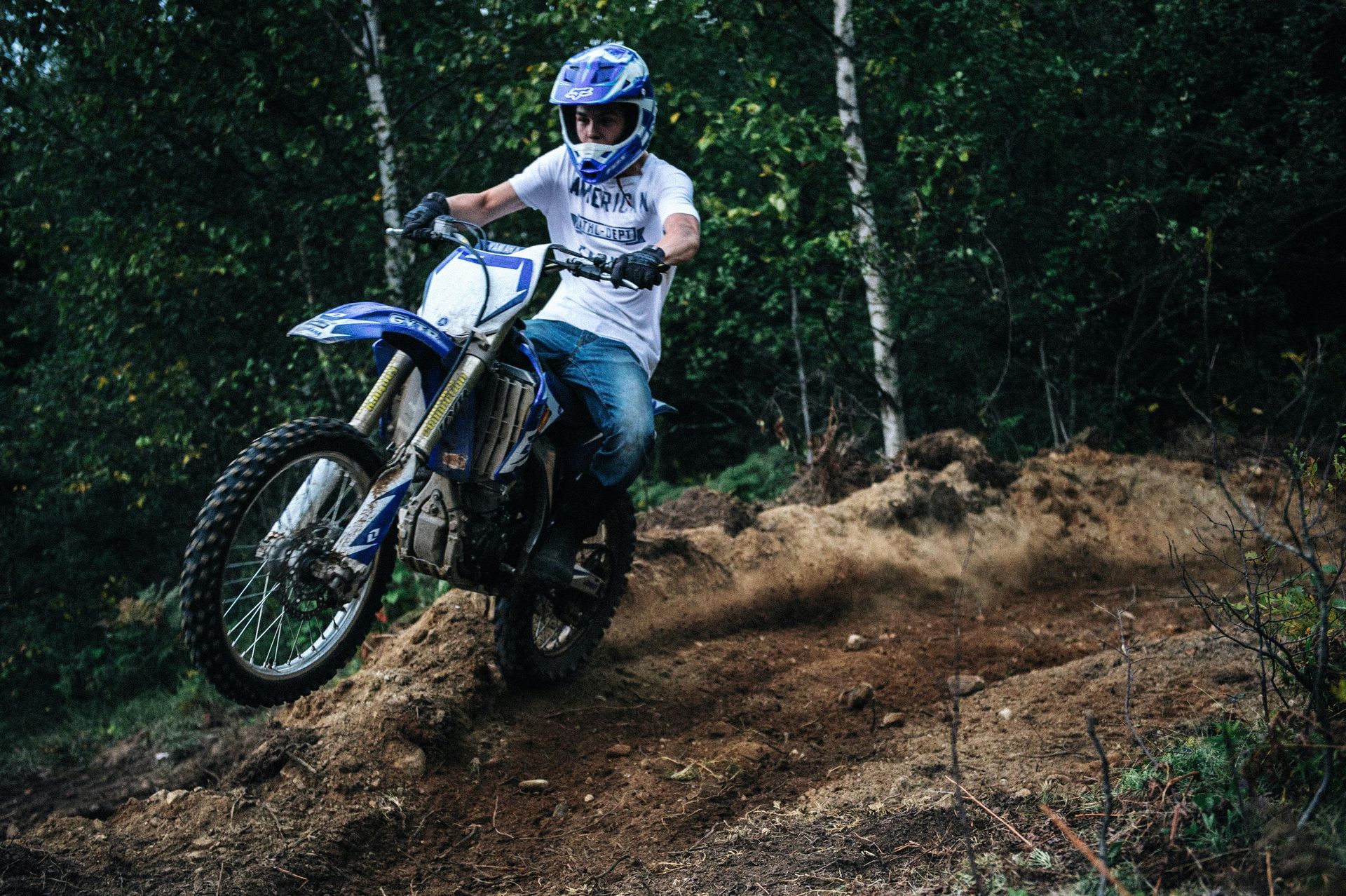 A person in a helmet riding a blue dirt bike over a dusty, wooded trail.