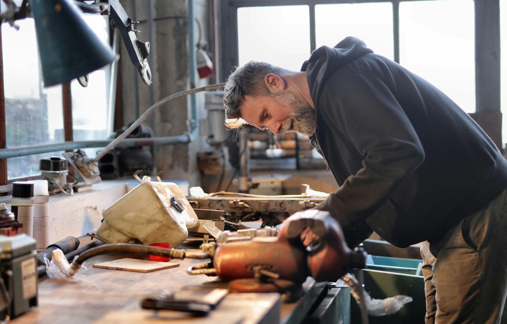 A focused person in a dark hoodie works at a cluttered workbench in a dimly lit, industrial workshop.
