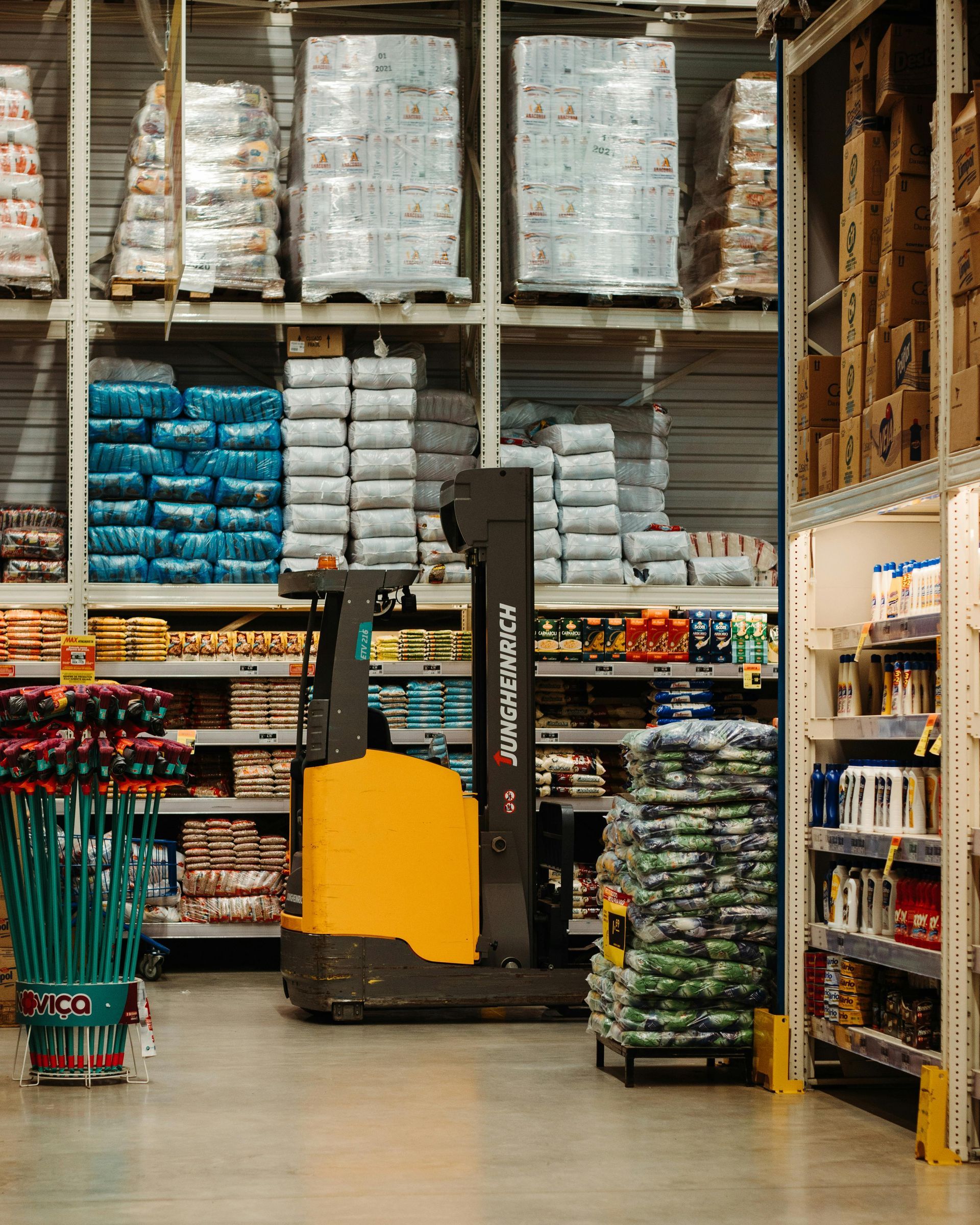 A long, narrow warehouse aisle flanked by tall orange industrial storage racks filled with boxes and pallets.
