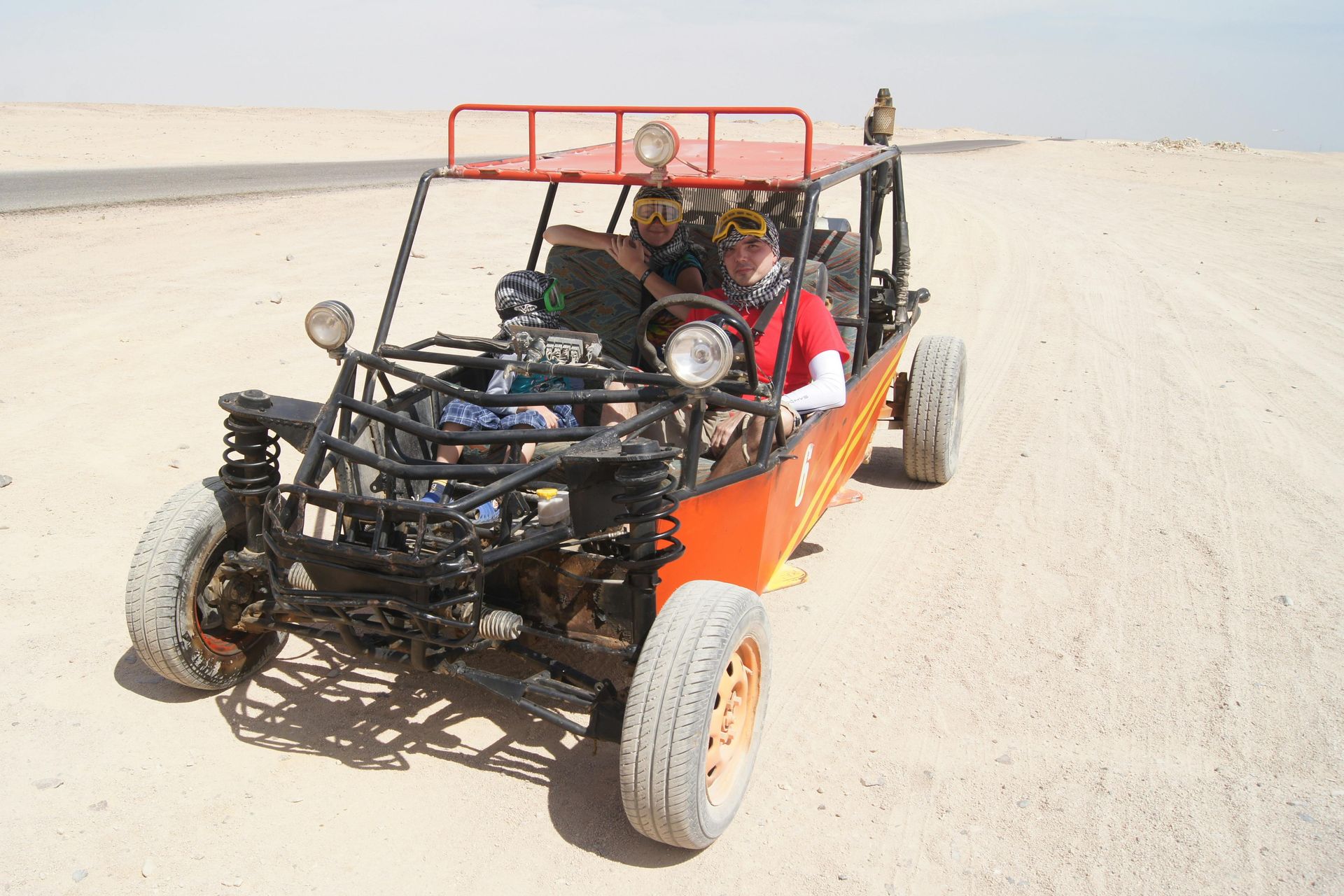 Two people in an orange dune buggy on a desert road with a sandy landscape.