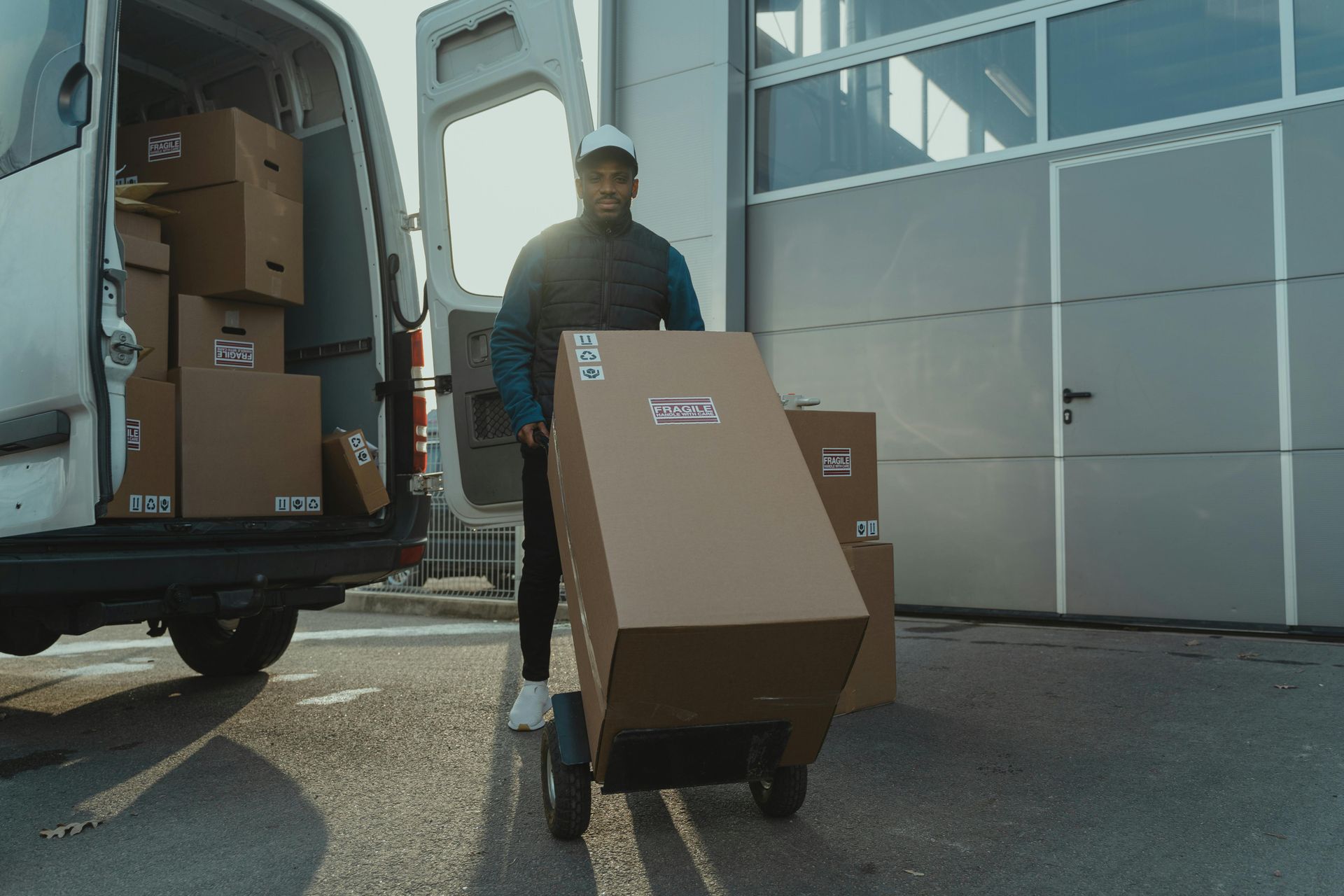 A person in a vest and hat pushes a tall cardboard box on a hand truck away from a delivery van in a paved lot.