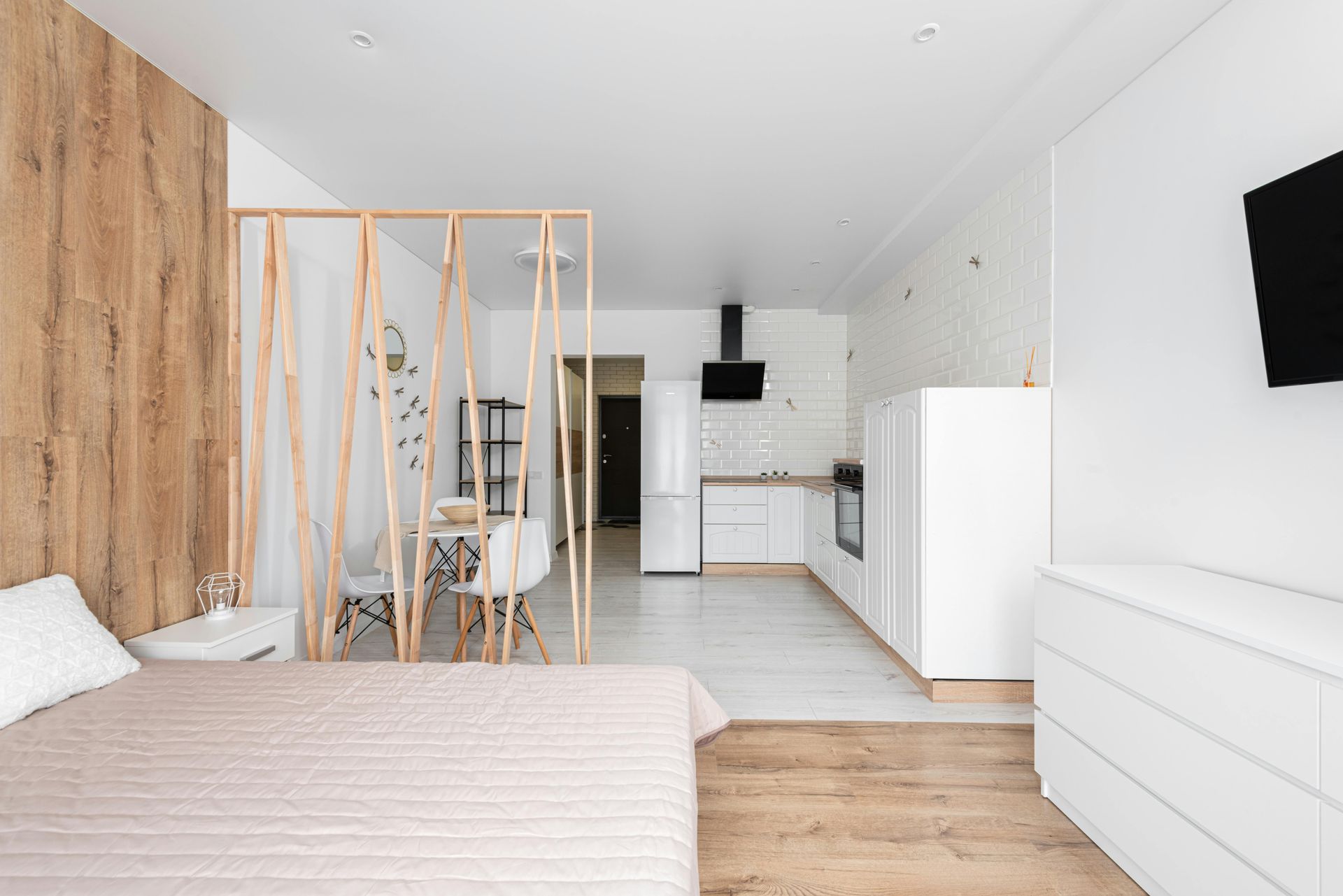 A bright studio apartment with a bed, a wood-slat room divider, a small dining area, and a white kitchen in the background.
