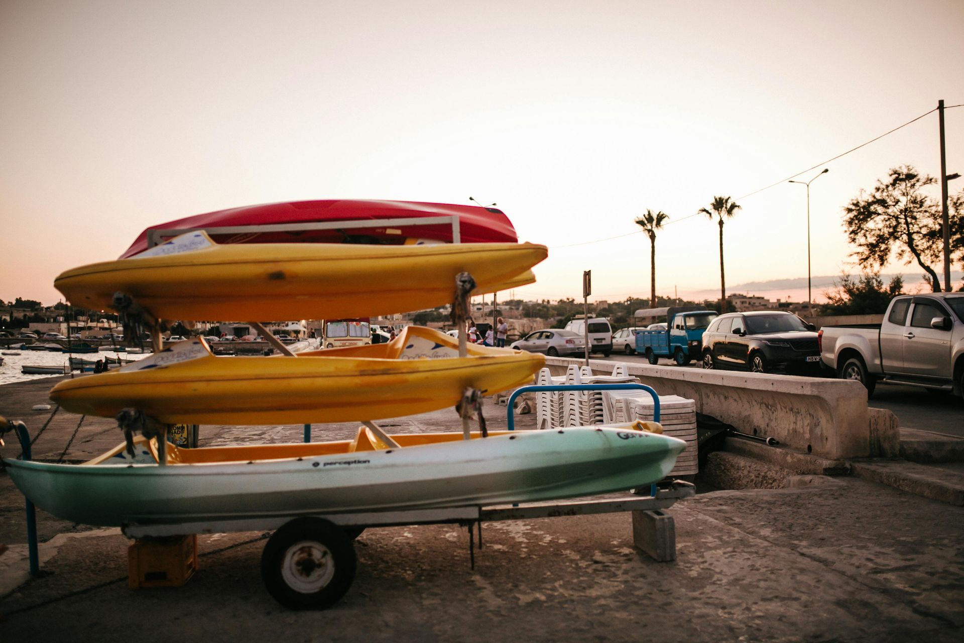 A trailer holds a stack of three kayaks—red, yellow, and pale green—parked on a concrete waterfront at sunset.