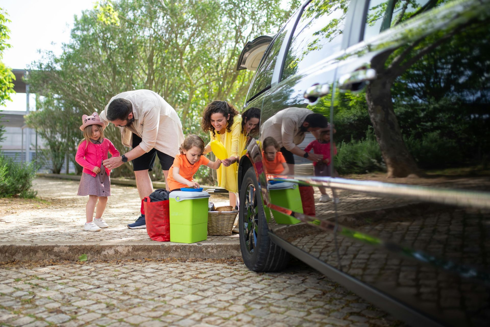 A family packs a black car for an outing, standing near luggage and a green cooler on a cobblestone driveway.