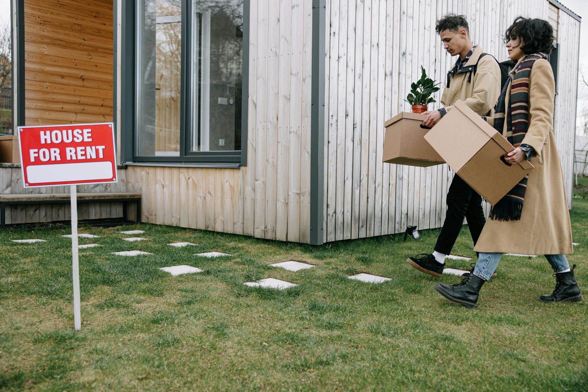 Two people carrying moving boxes toward a modern house with a