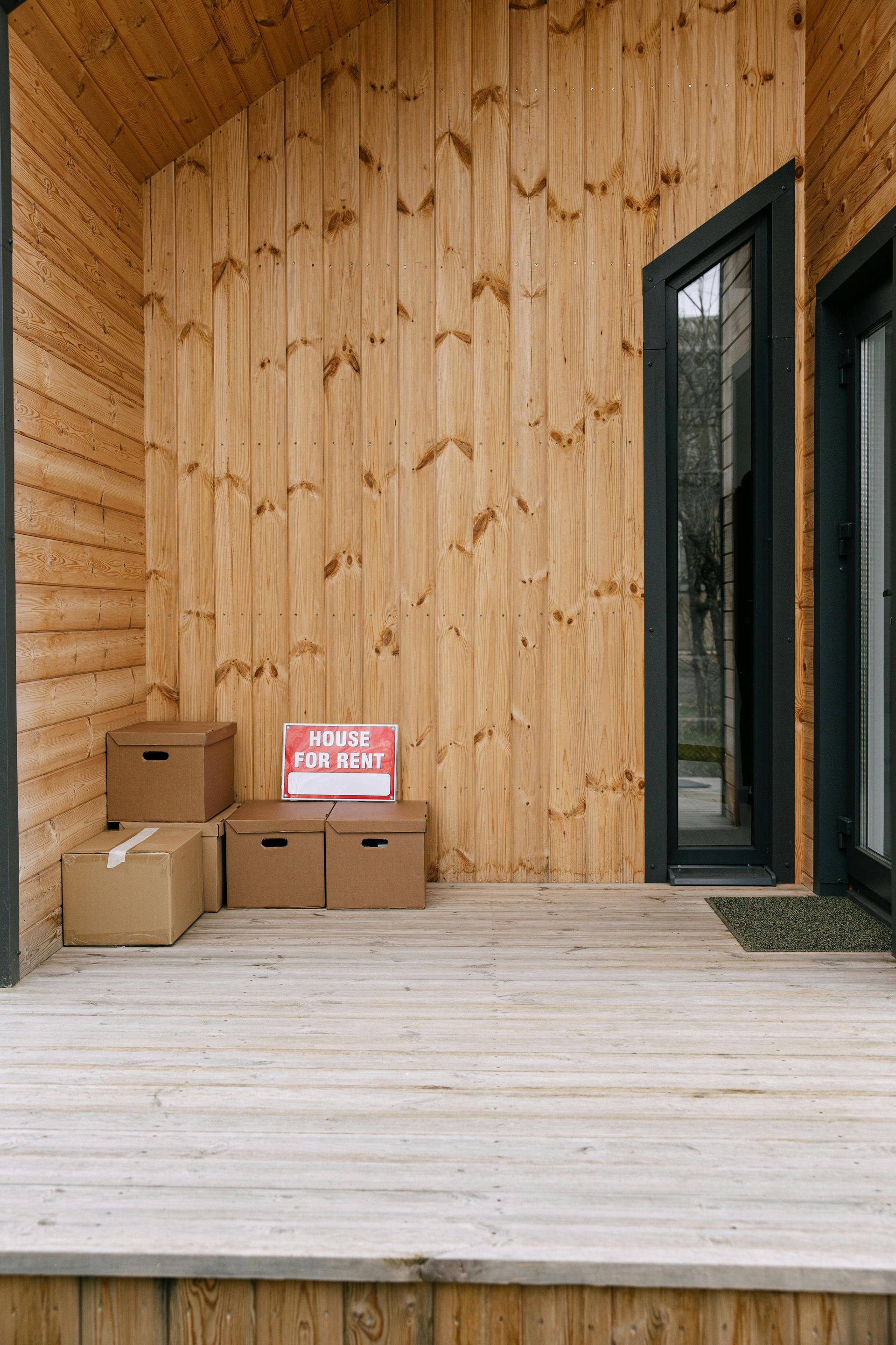 Three cardboard boxes and a small sign sit on a wooden porch outside a building with light wood paneling and black doors.