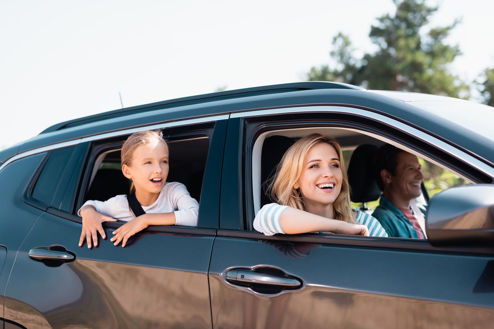 A child and two adults smiling while leaning out of the open windows of a dark car on a sunny day.