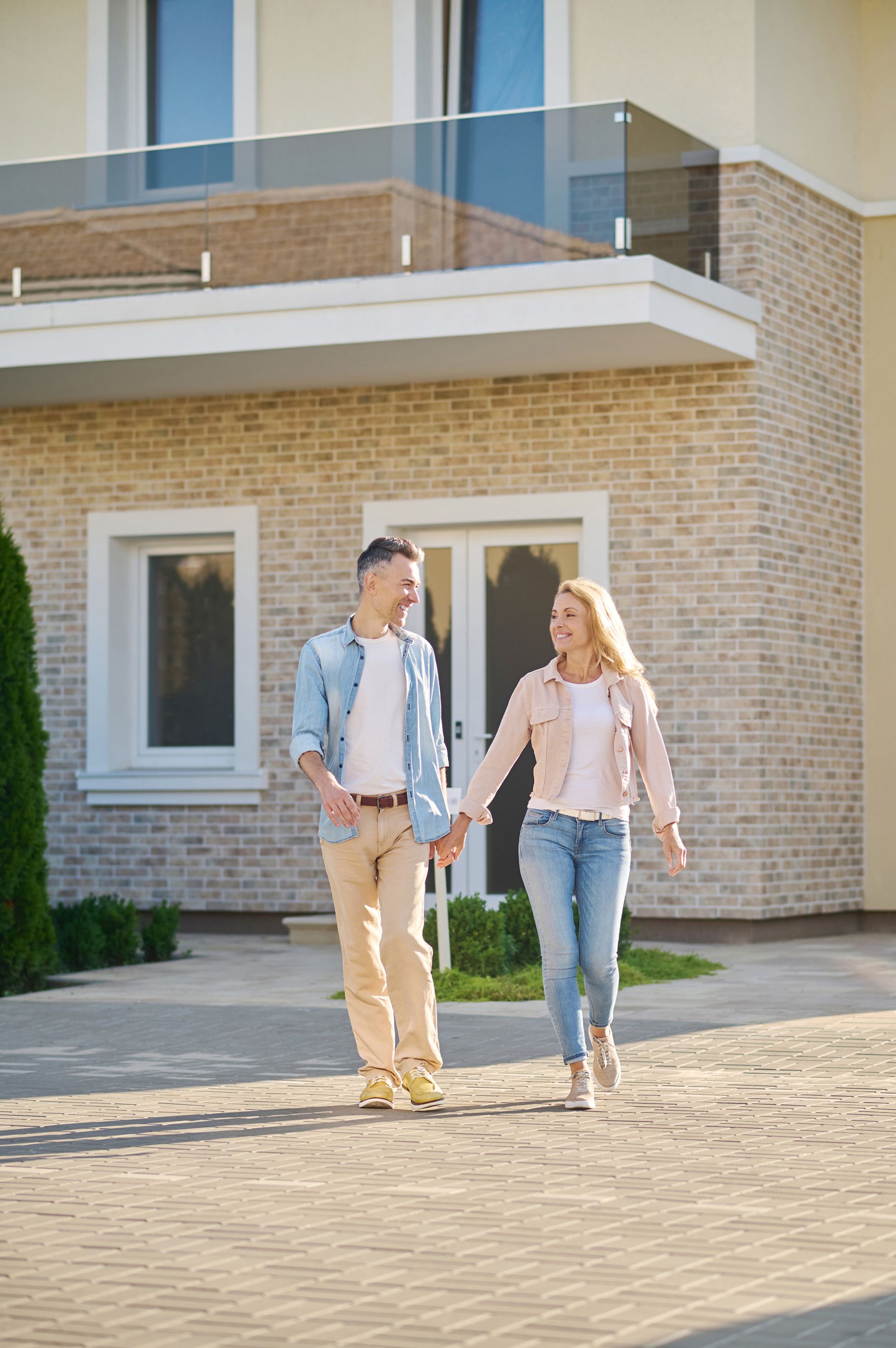 A couple walks hand-in-hand toward the camera in front of a modern house with brick walls and a glass-railed balcony.