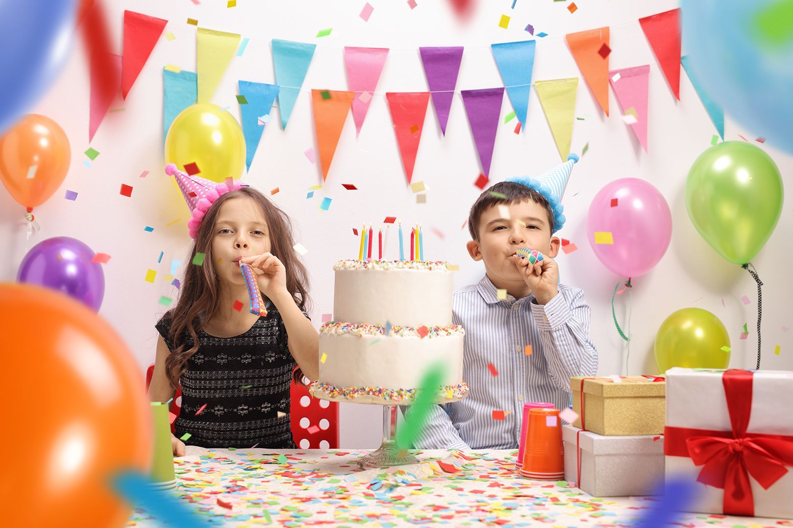 Two children blowing party horns at a birthday table with cake, balloons, gifts, and colorful decorations