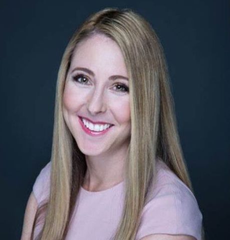 A woman with long blonde hair is smiling and wearing a pink shirt.