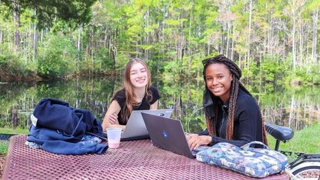 Two women are sitting at a picnic table with laptops.