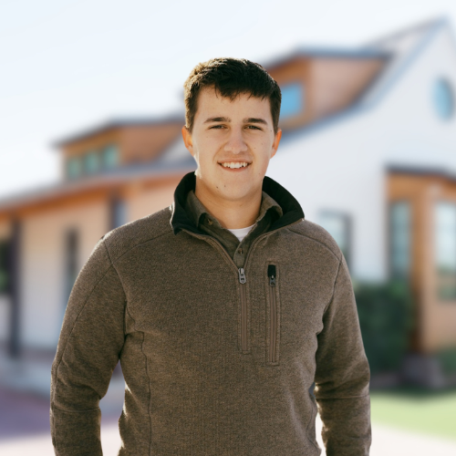 A man in a brown sweater stands in front of a house