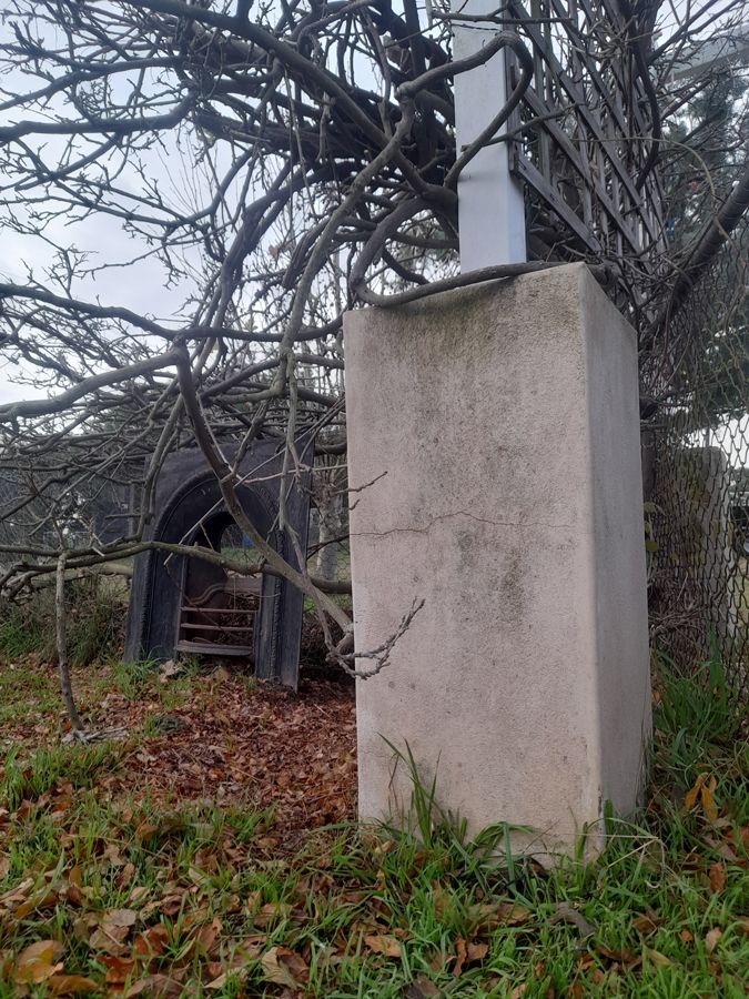 Concrete pillar with a cross, trees, and an arched structure in the background.