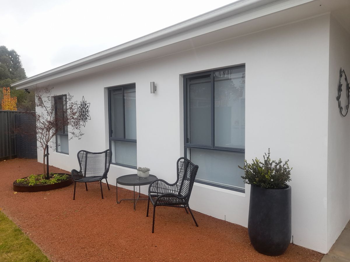 White building with black trim, two windows with white curtains, pink flowers in front.