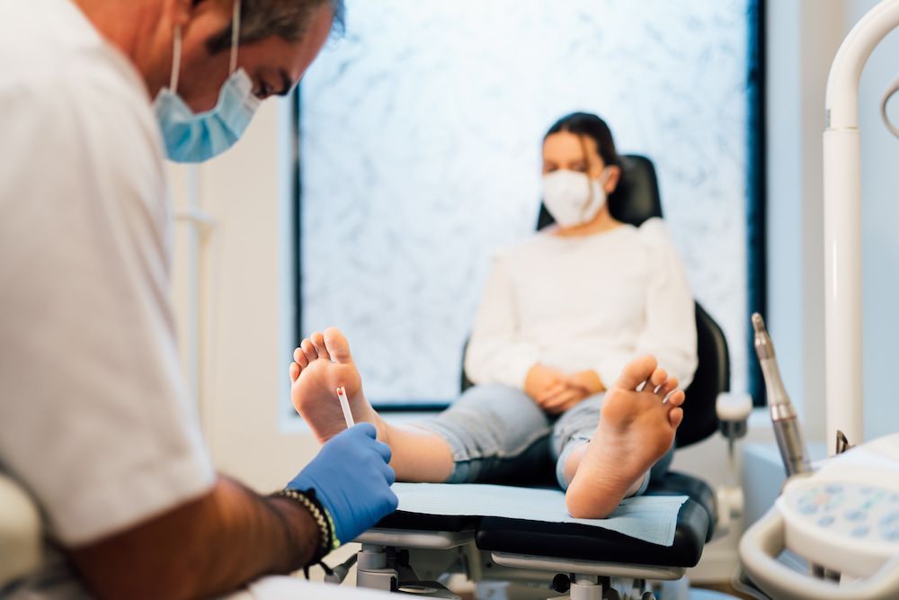 A Woman Is Sitting In A Chair Getting Her Feet Examined By A Doctor — Lake Macquarie Podiatry in Swansea, NSW