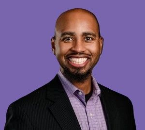 A man in a suit and purple shirt is smiling in front of a purple background.