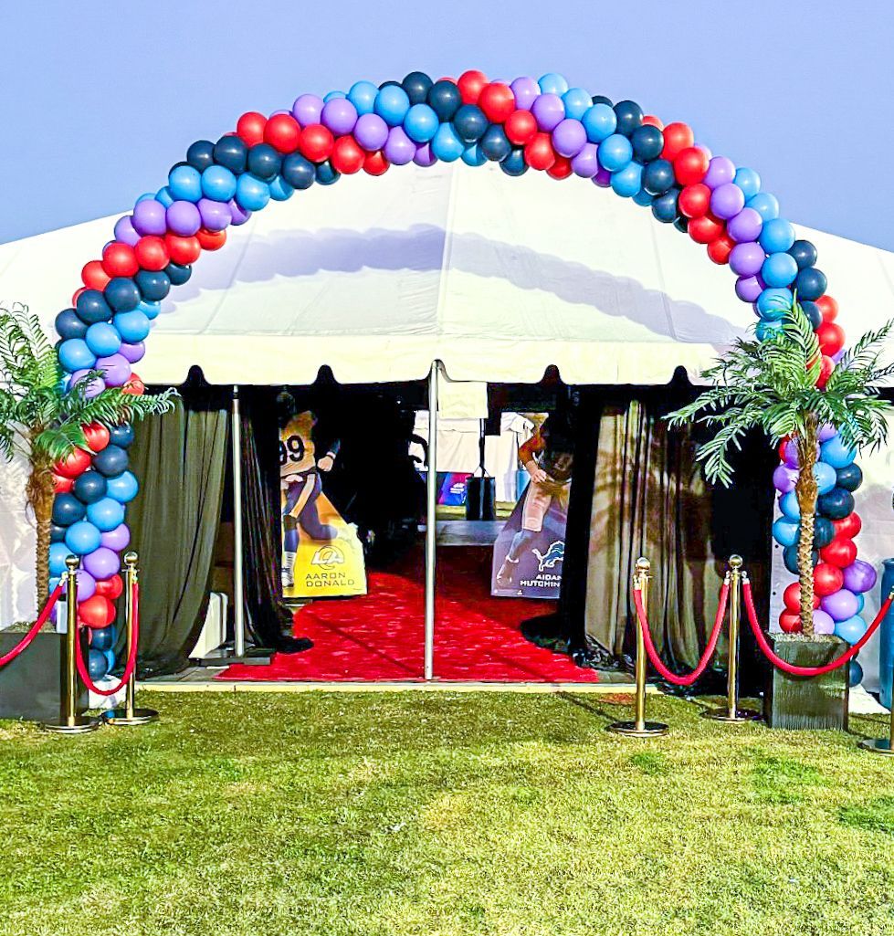 A tent entrance with a red carpet and a balloon arch in blue, red, and purple, with palm tree decorations.