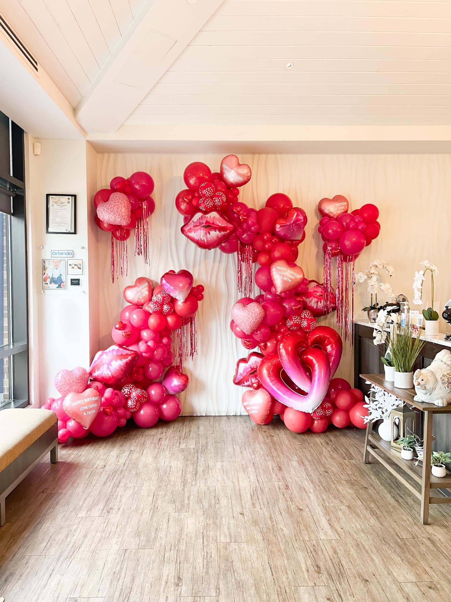 Red and pink heart-shaped balloon display against a white wall with fringe. A side table with flowers sits to the right.