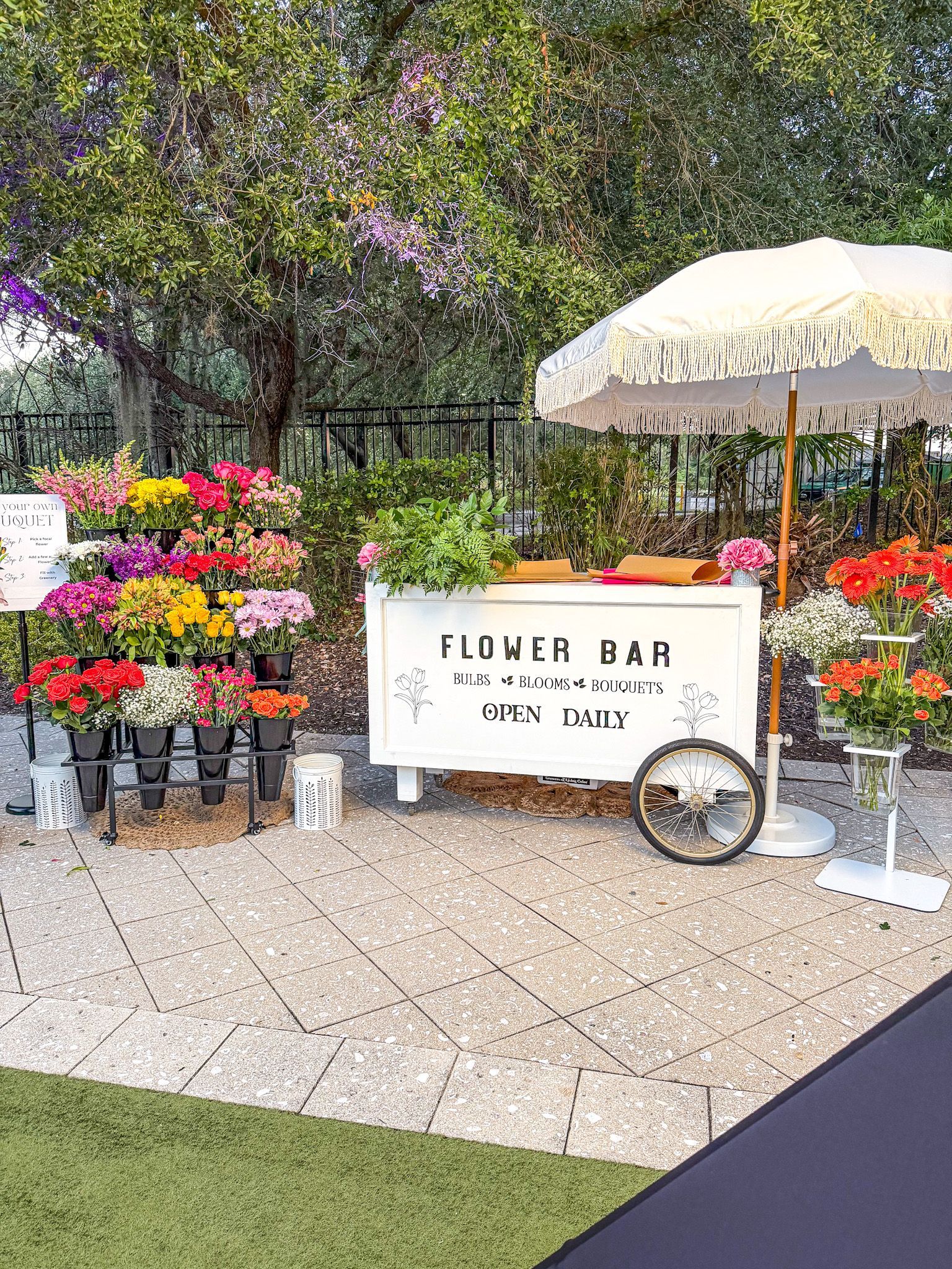 Flower bar with colorful flowers, white cart, umbrella, and a paved outdoor setting.