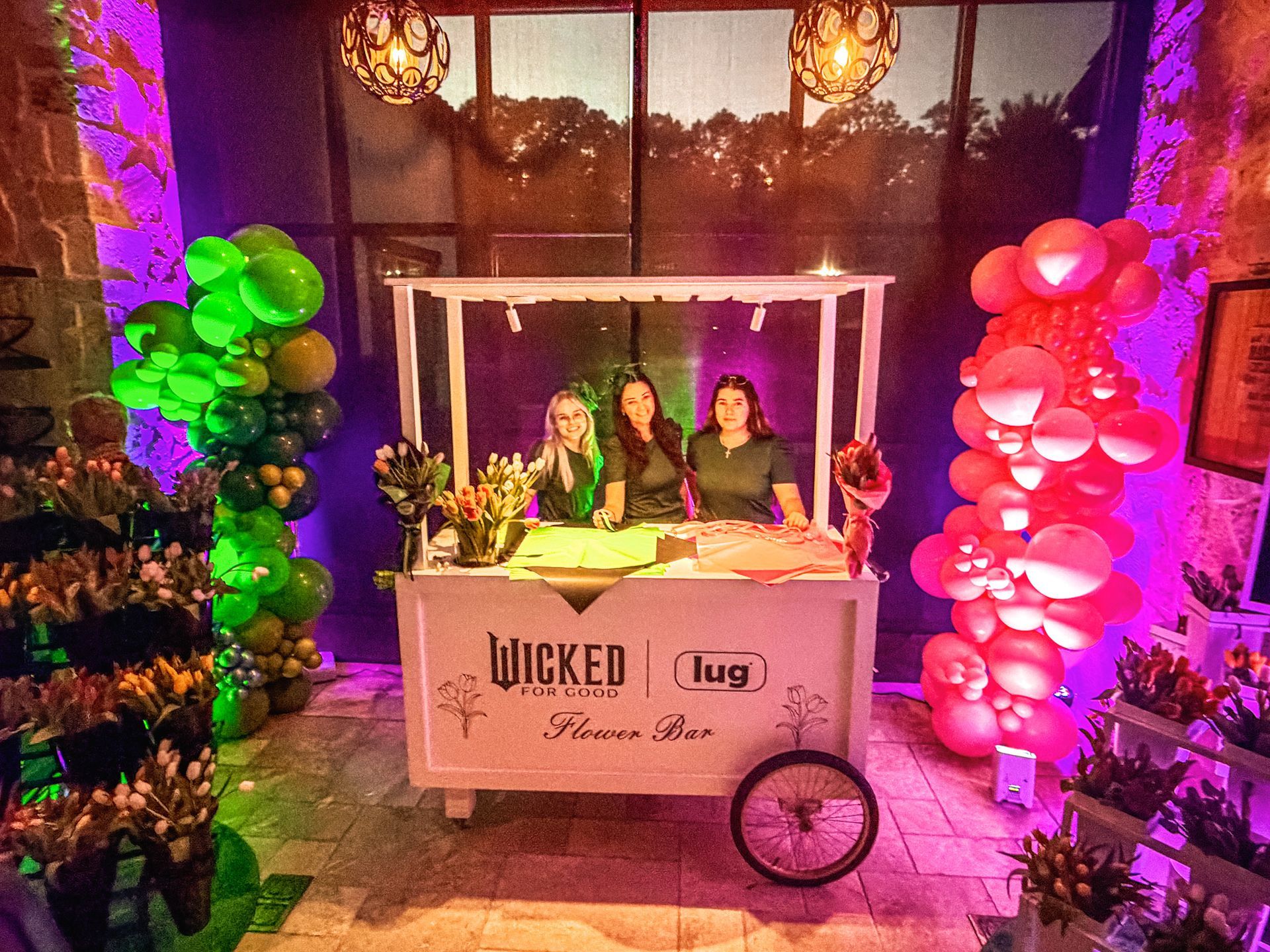 Flower bar with three people, green and pink balloon arches, and flower displays.