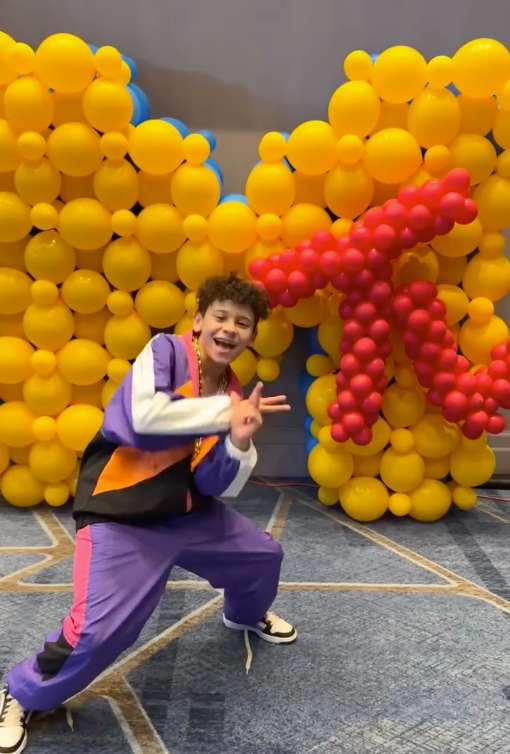 A boy is standing in front of a wall of yellow and red balloons