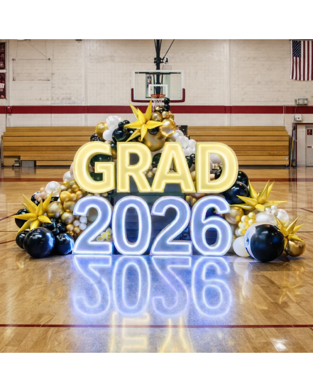 Graduation celebration setup with GRAD 2026 in neon lights, surrounded by black, gold, and white balloons, in a gymnasium.