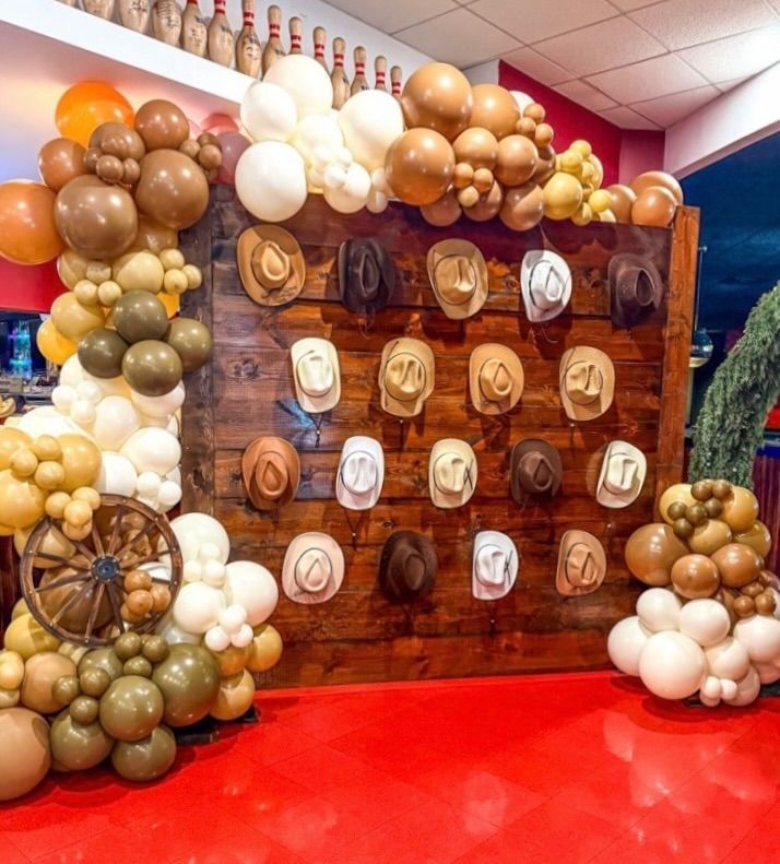 Western-themed photo booth with cowboy hats on wooden backdrop, decorated with balloons in earth tones.