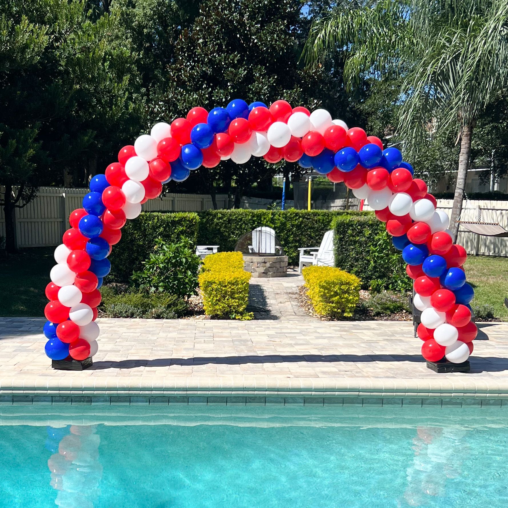 A red , white and blue balloon arch is sitting next to a pool.