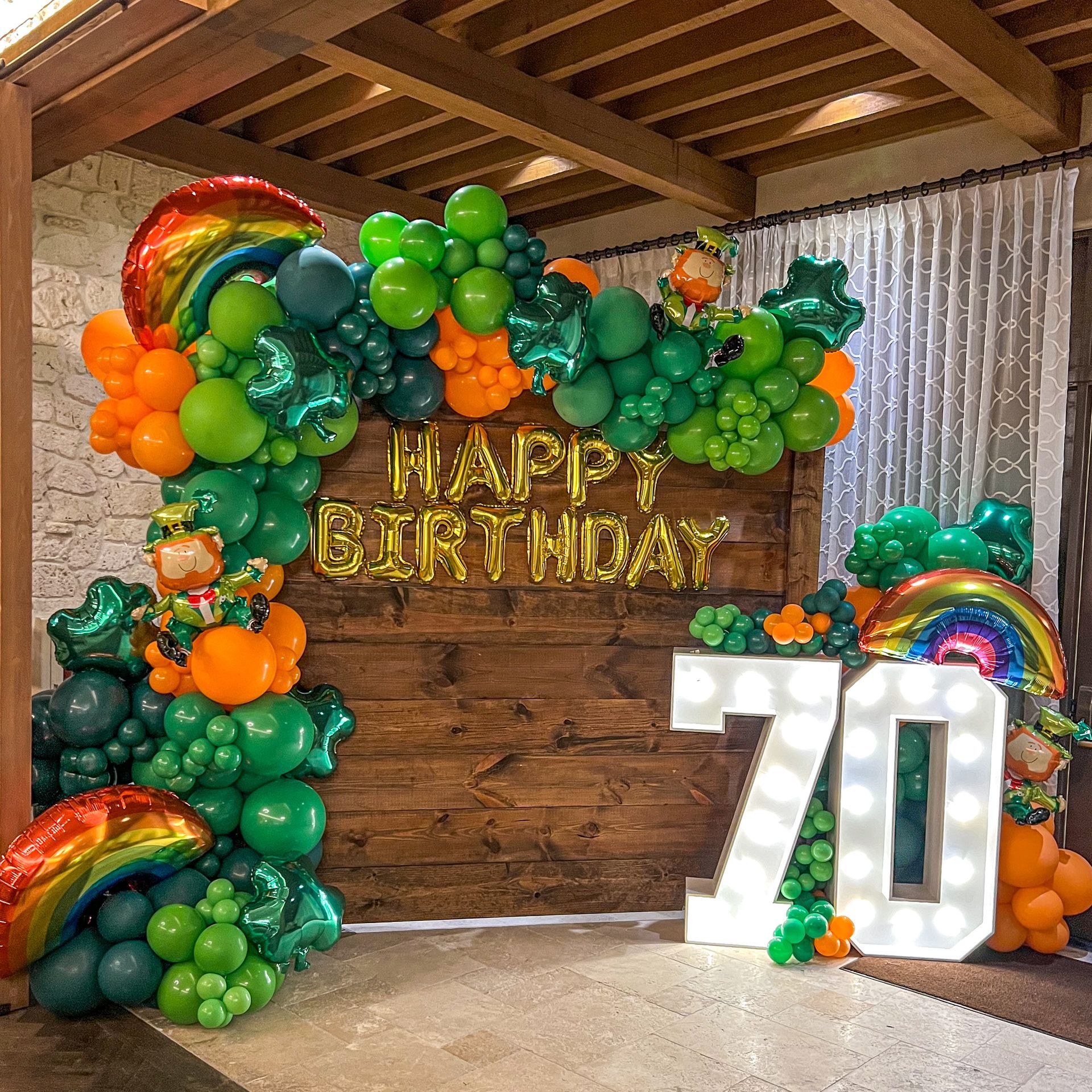 A wooden wall is decorated with green and orange balloons for a 70th birthday.