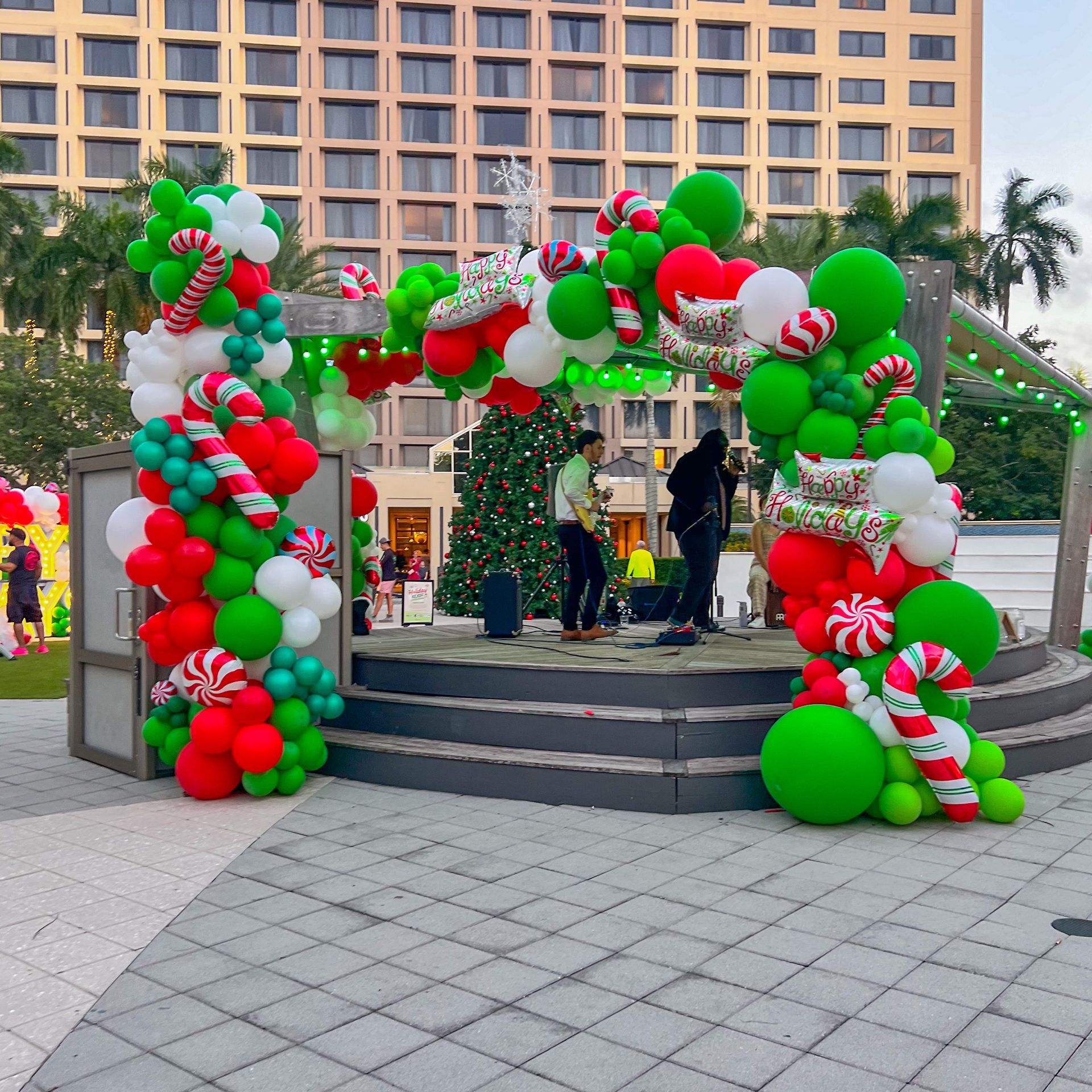 Christmas arch of red, green, and white balloons frames a stage with musicians; Christmas tree and building in background.