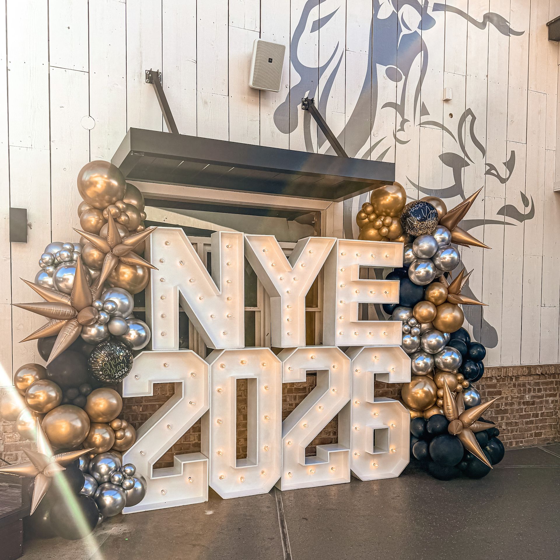 NYE 2026 sign with lit-up letters, decorated with gold, silver, and black balloons and starbursts, near a building.