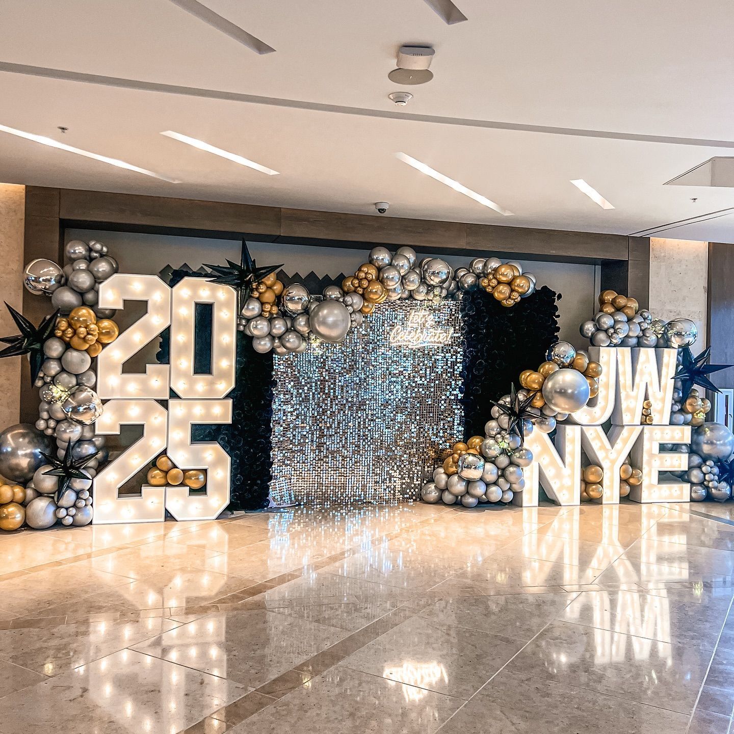 New Year's Eve party decorations with large illuminated numbers 2025 and JW NYE. Balloons in silver, gold, and black.
