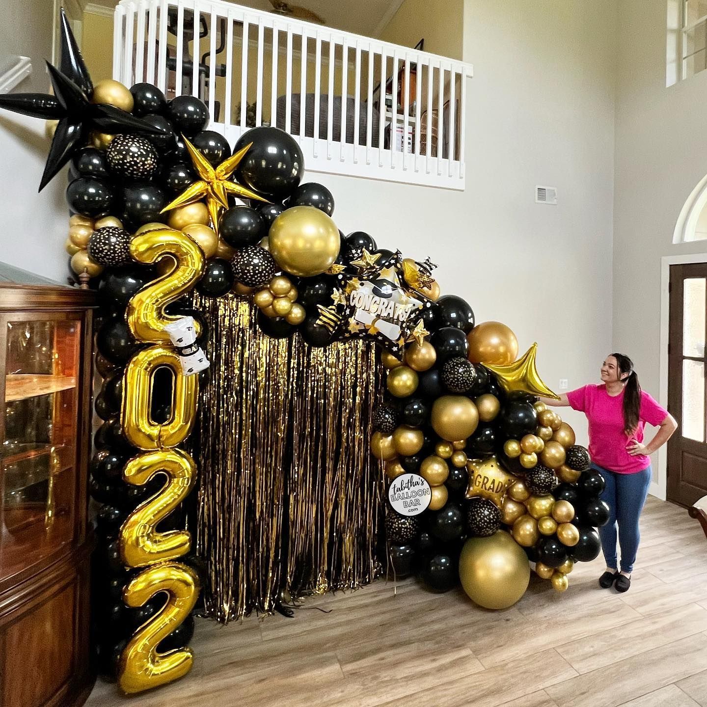A woman is standing in front of a wall decorated with black and gold balloons.