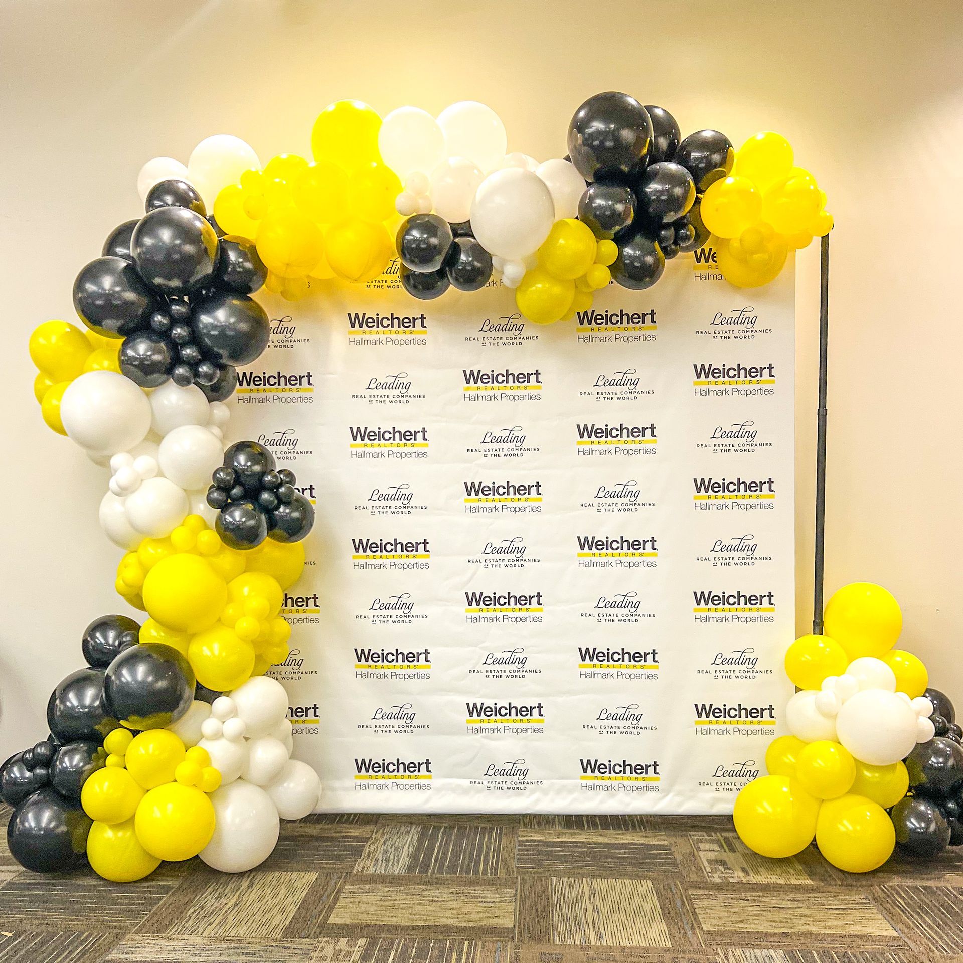 Balloon arch in black, yellow, and white colors in front of a white backdrop with logo.