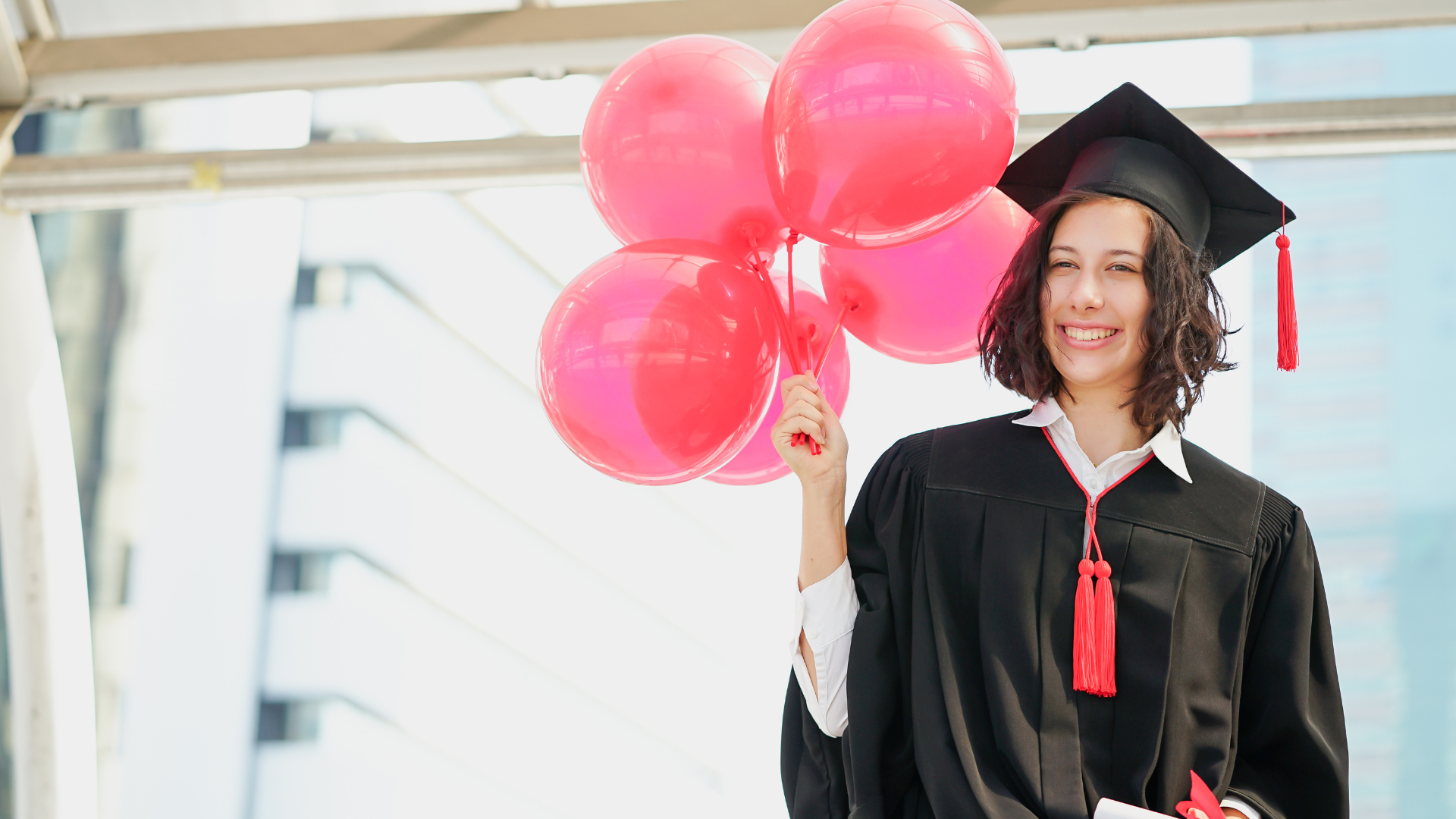 A woman in a graduation cap and gown is holding a bunch of pink balloons.