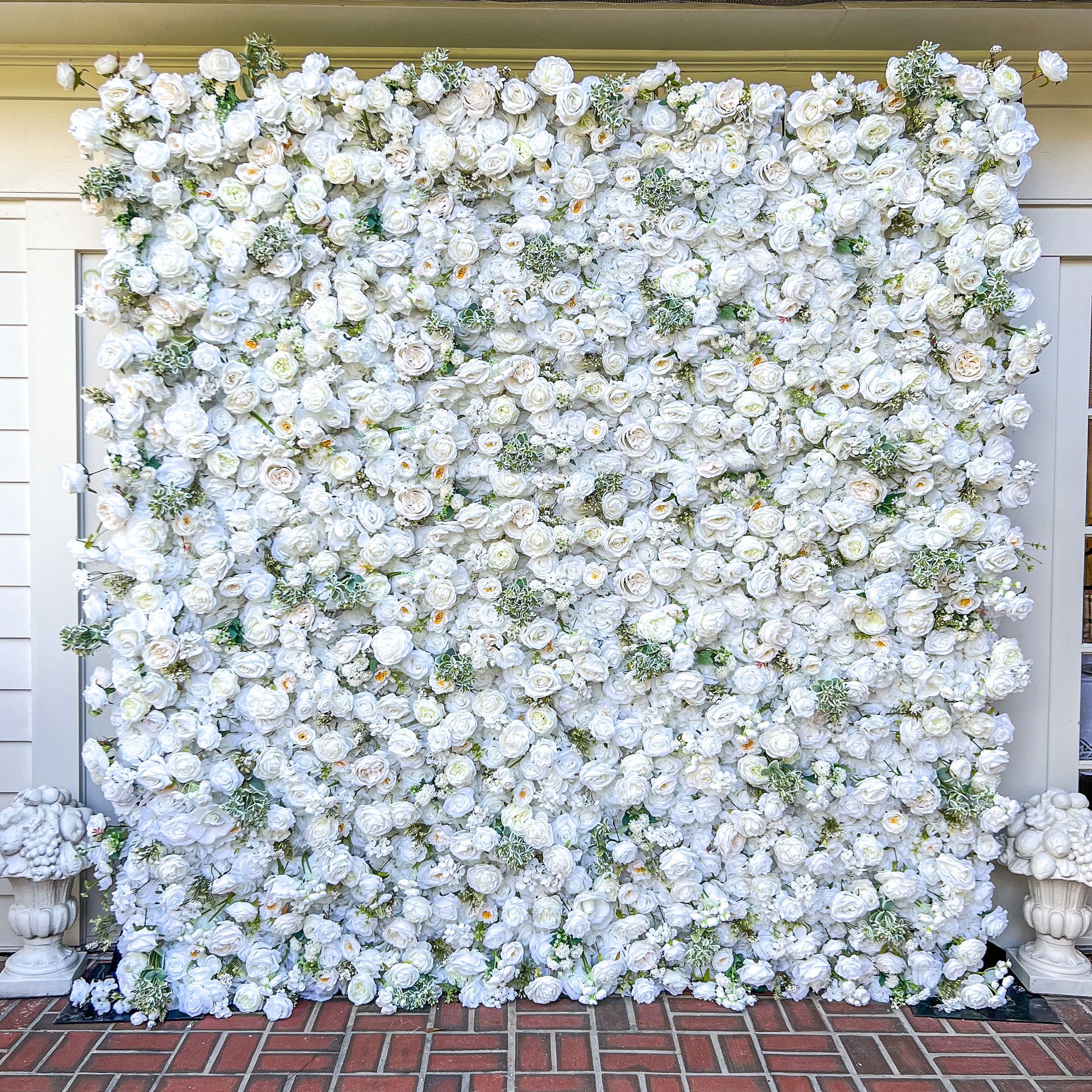 White floral wall backdrop with accents of greenery, set against a building with brick flooring.