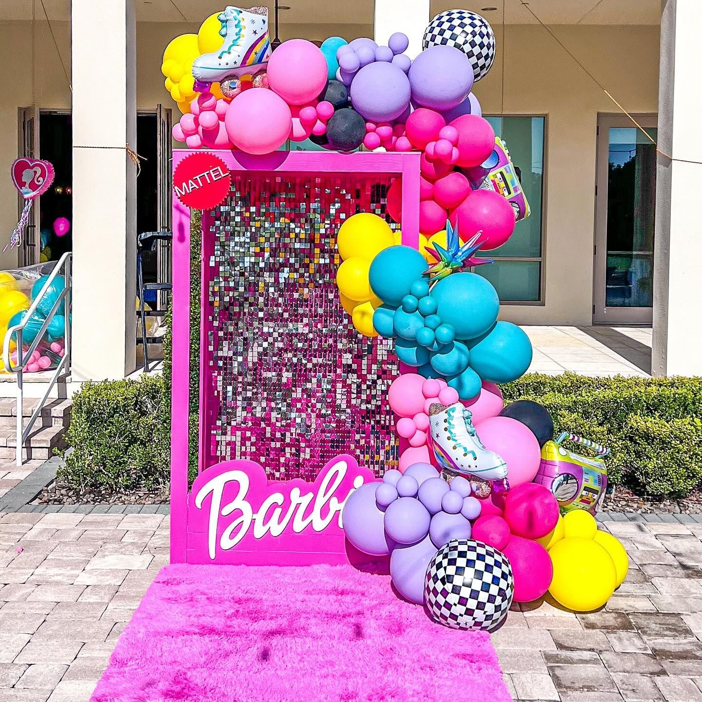 A barbie box is surrounded by balloons and a pink carpet.