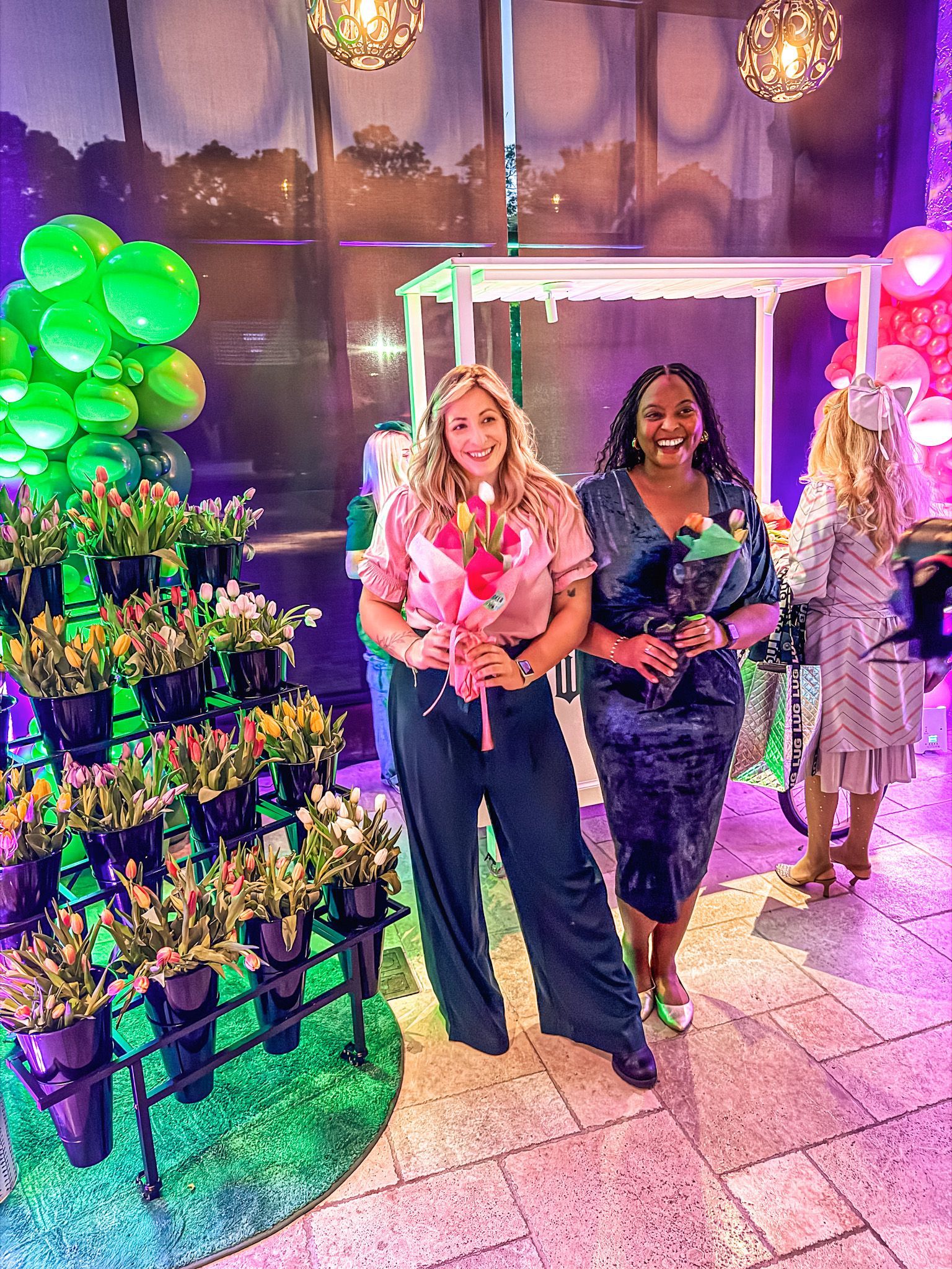 Two women hold flowers, pose near floral display, balloons, under decorative lights.