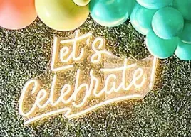 a neon sign that says `` let 's celebrate '' is sitting on a carpet next to balloons