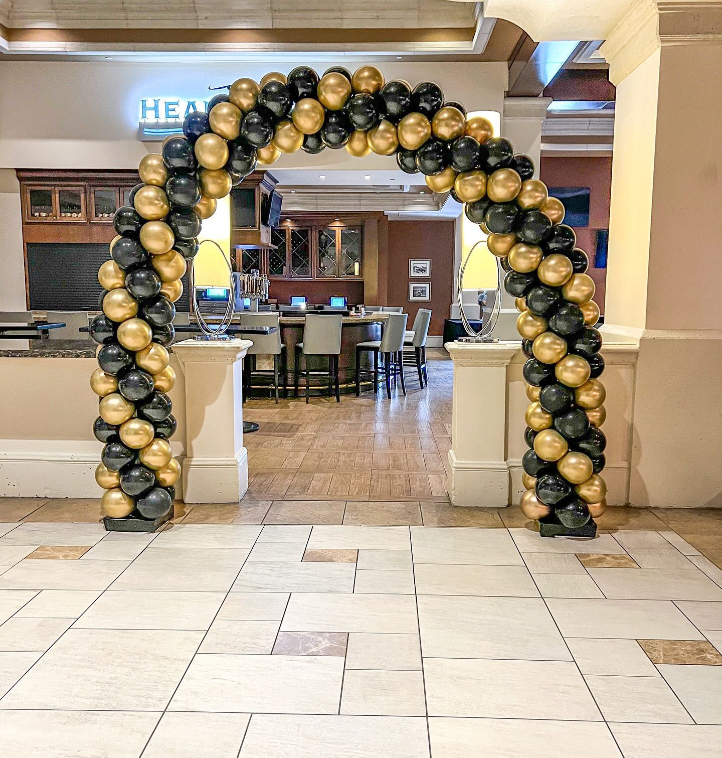 Black and gold balloon archway in an entryway, leading to a room with bar and tables.