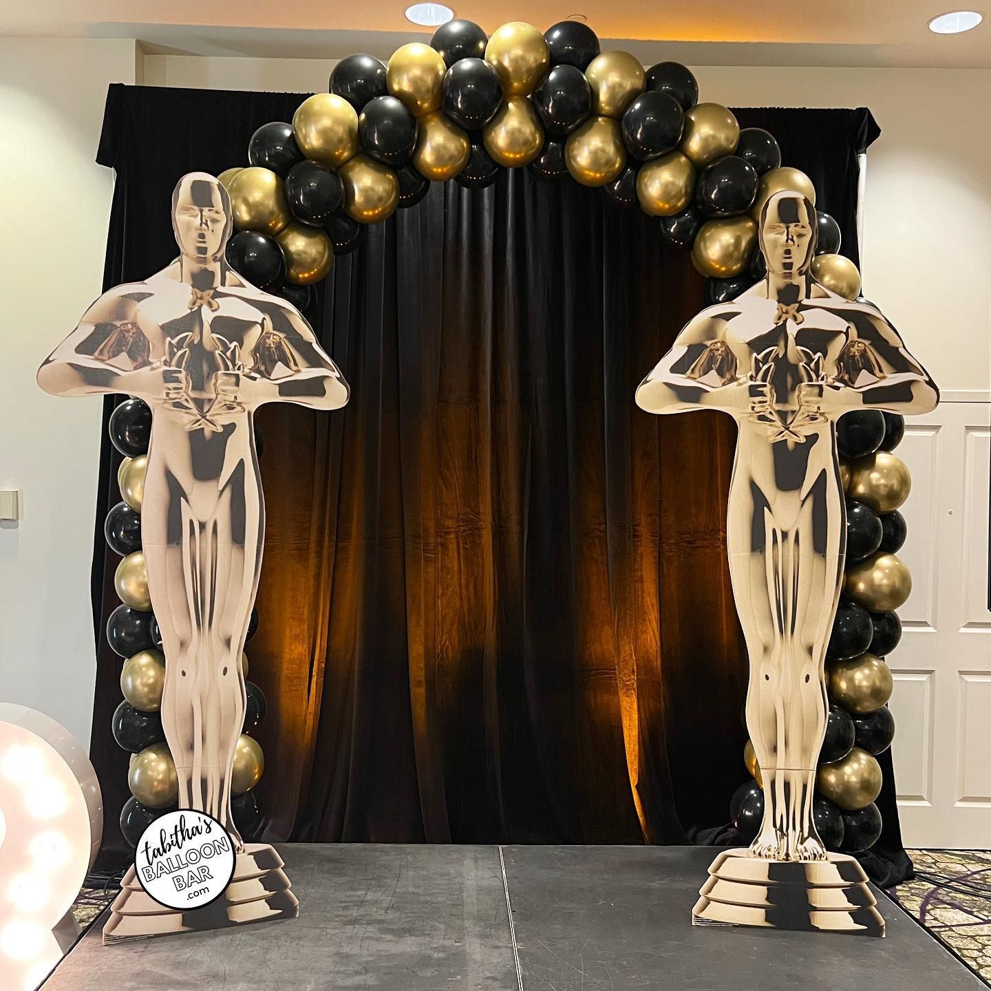 Two oscar statues are standing in front of a black and gold balloon arch.