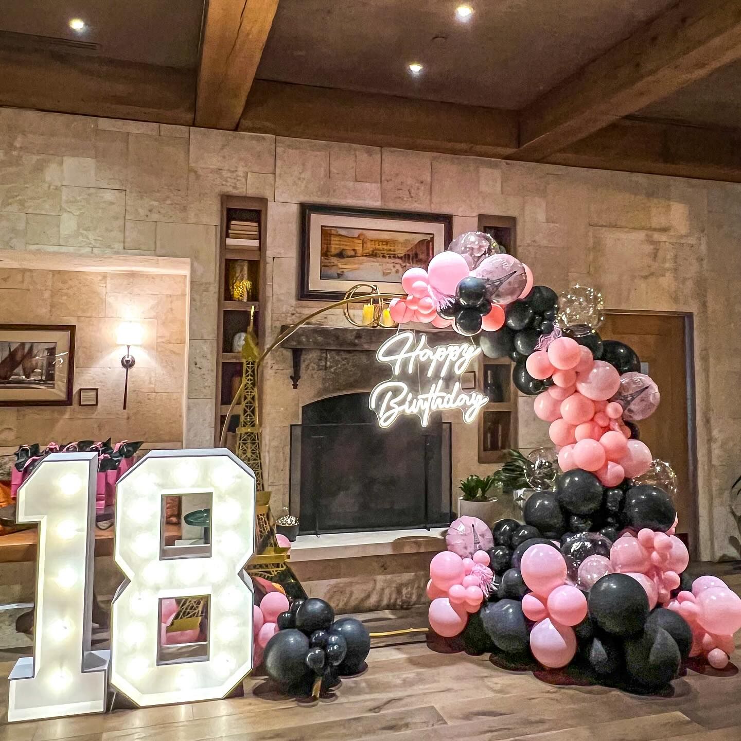 A room decorated with pink and black balloons and a neon sign that says happy birthday.