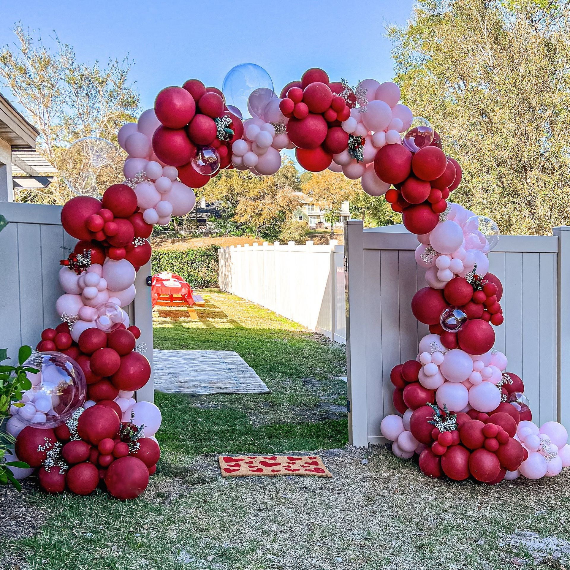 A red and pink balloon arch is sitting in front of a white fence.