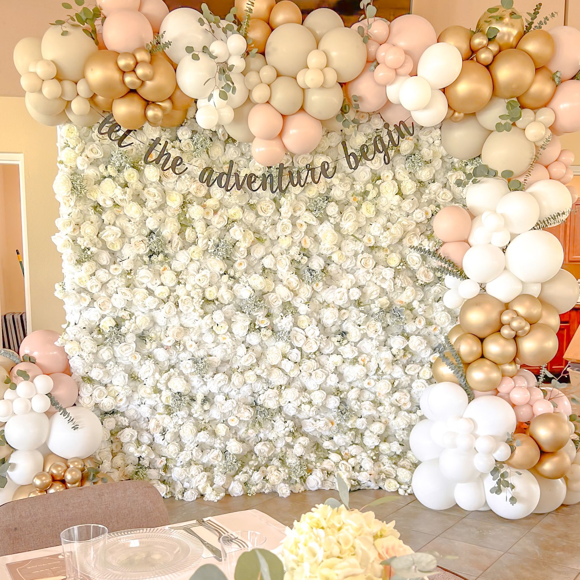 A wall of flowers and balloons with a sign that says `` let the adventure begin ''