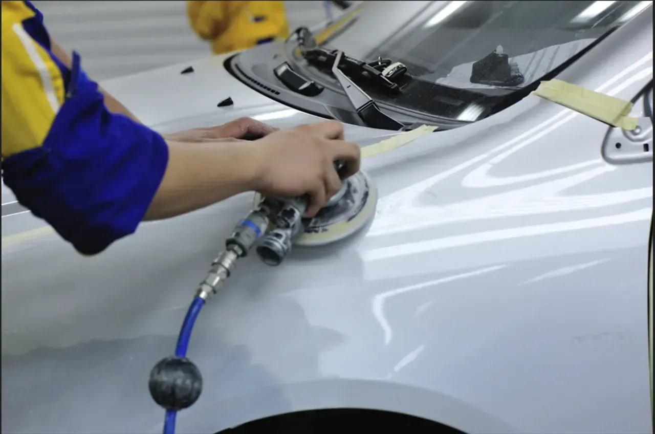 Person using a sander on a car's painted surface, likely for detailing or repair. Blue and yellow work clothing.