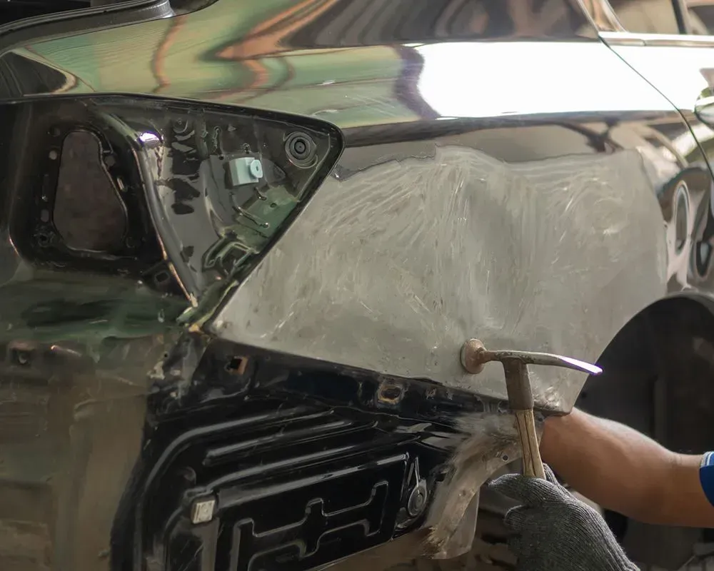 Car body repair, person hammering a dented black car panel with gray filler.