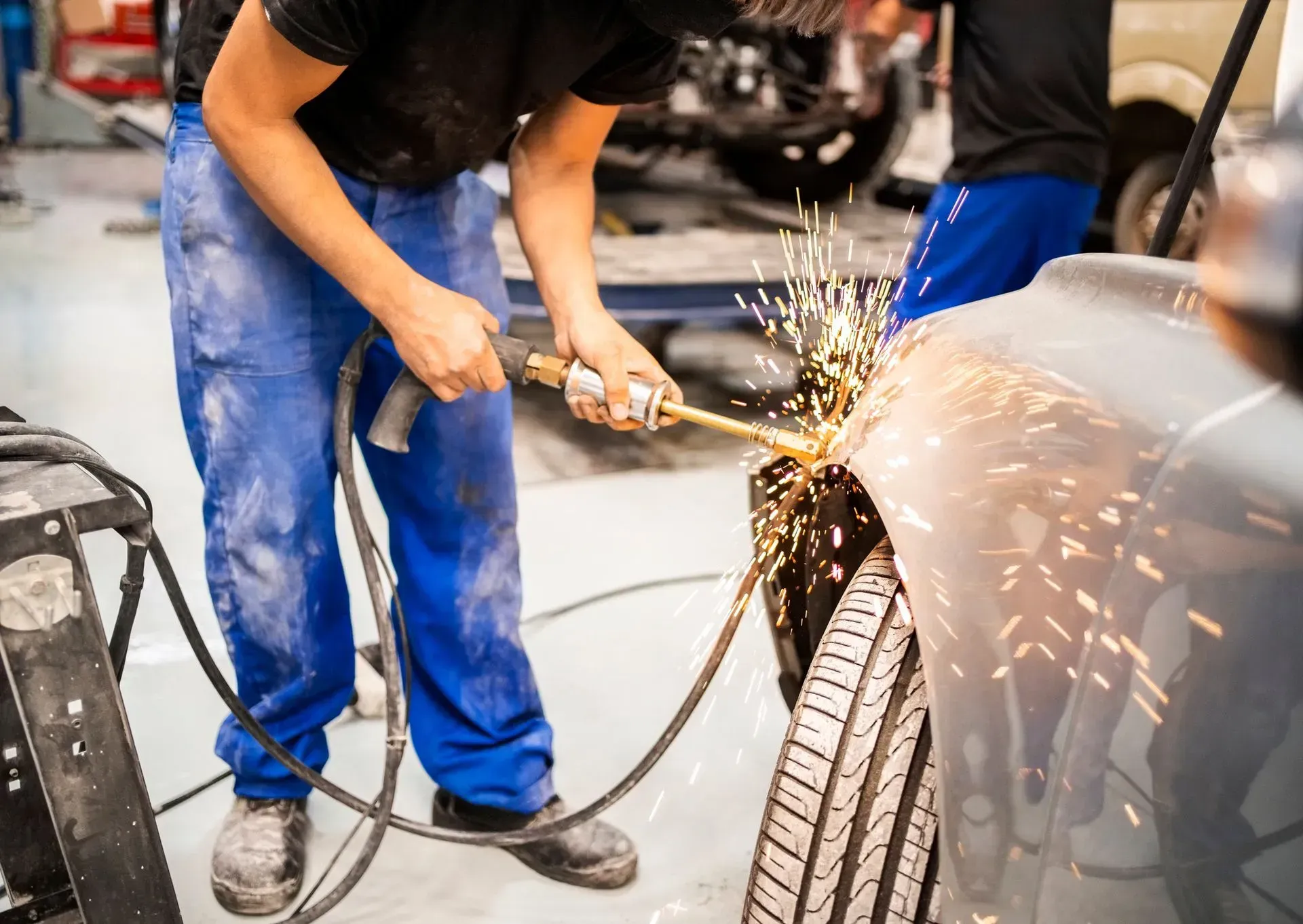Mechanic using a welding torch on a car fender, creating sparks in a repair shop.